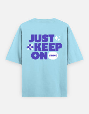 Oversized t-shirt with bold purple typography reading “Just Keep On Vibing,” decorated with a flower and sparkle icons for a retro-positive vibe.
